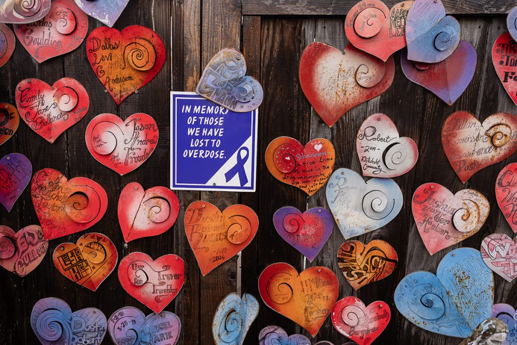 Metal hearts with the names of people in Nanaimo who have died from an overdose are affixed to a wodden fence with a sign reading "In memory of those we have lost to overdose."