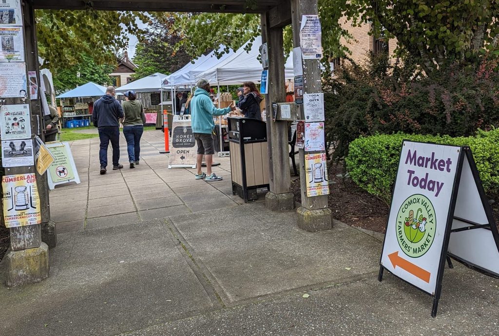 A sign outside the Cumberland Farmers' Market that says 'market today,' and people walk around booths set up in the background.