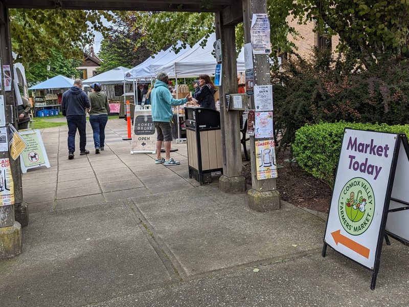 A sign outside the Cumberland Farmers' Market that says 'market today,' and people walk around booths set up in the background.