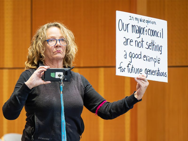 This image has an empty alt attribute; its file name is 2025.02.24-women-with-sign-and-phone-at-Nanaimo-city-council-meeting-by-Mick-Sweetman-1024x683.jpg A woman holds a protest sign and a cell phone during a Nanaimo city council meeting.