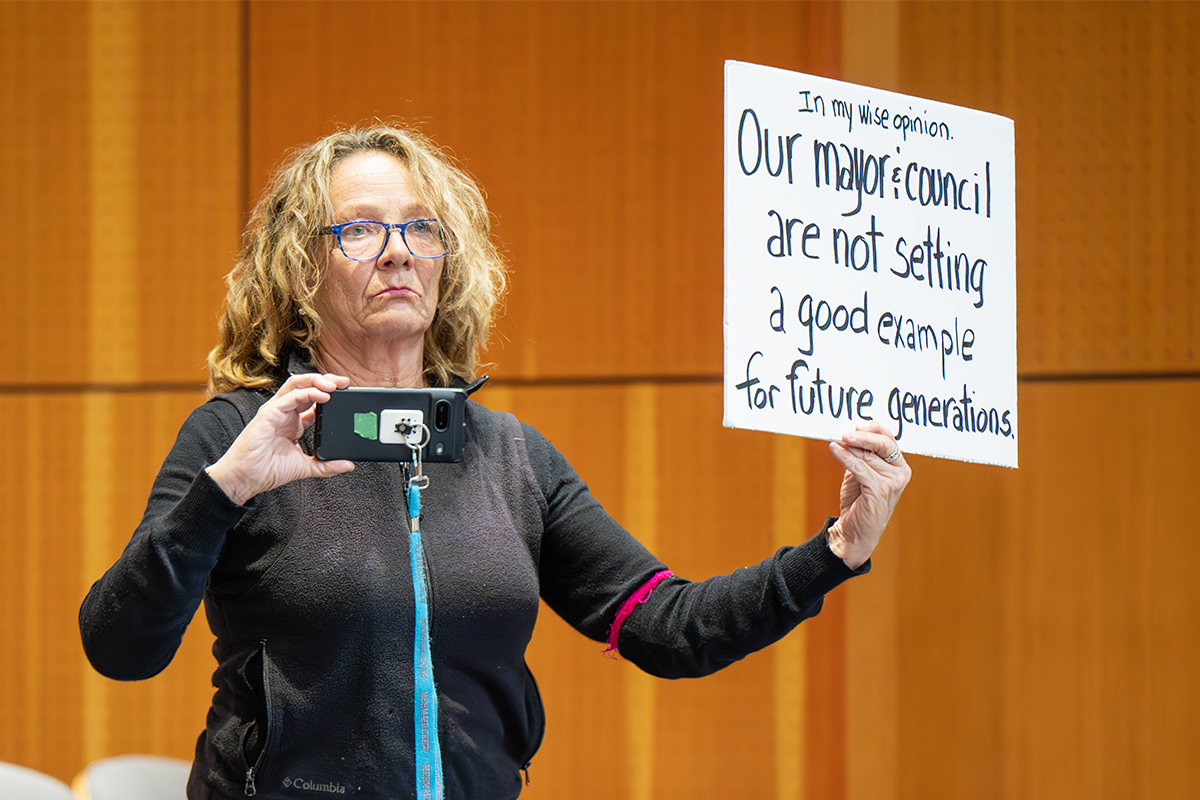 This image has an empty alt attribute; its file name is 2025.02.24-women-with-sign-and-phone-at-Nanaimo-city-council-meeting-by-Mick-Sweetman-1024x683.jpg A woman holds a protest sign and a cell phone during a Nanaimo city council meeting.