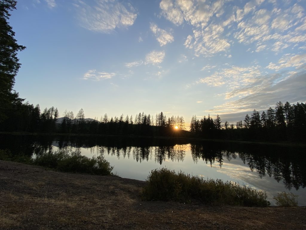The sun sets behind forest and hills in the background with a glassy lake in the foreground. Taken on Vancouver Island.