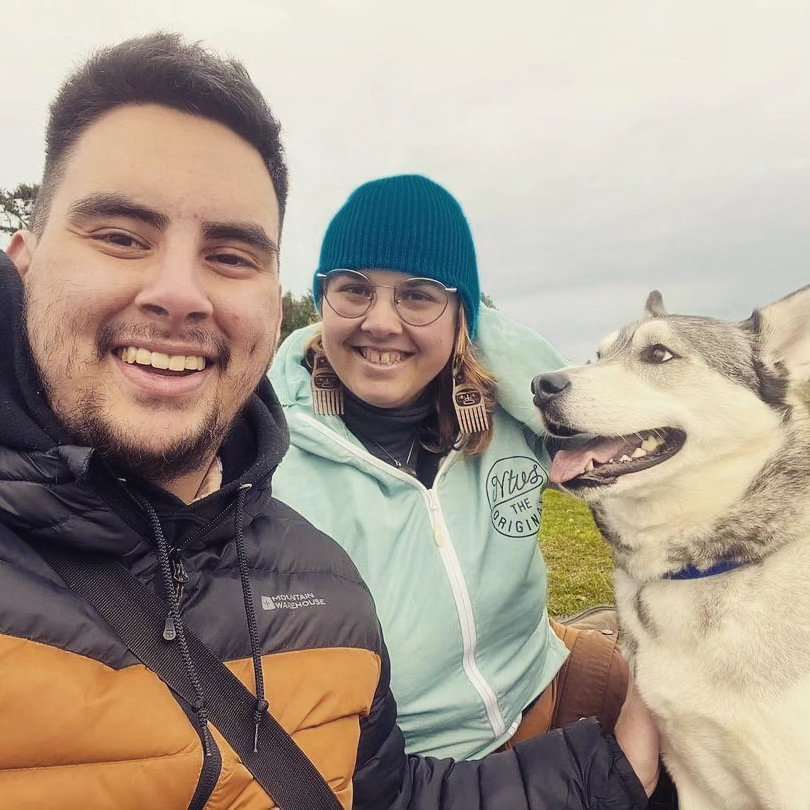 Two people outside smiling with a husky dog to the right. It's Jesse and Raini.