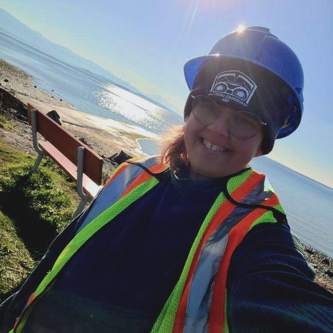 A person, Raini, near a beach takes a selfie with a hard hat and a high viz vest on.