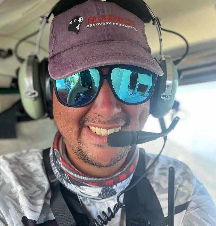 A man sits in a helicopter with headphones on and sunglasses. His hat reads 'Marmot Recovery foundation'