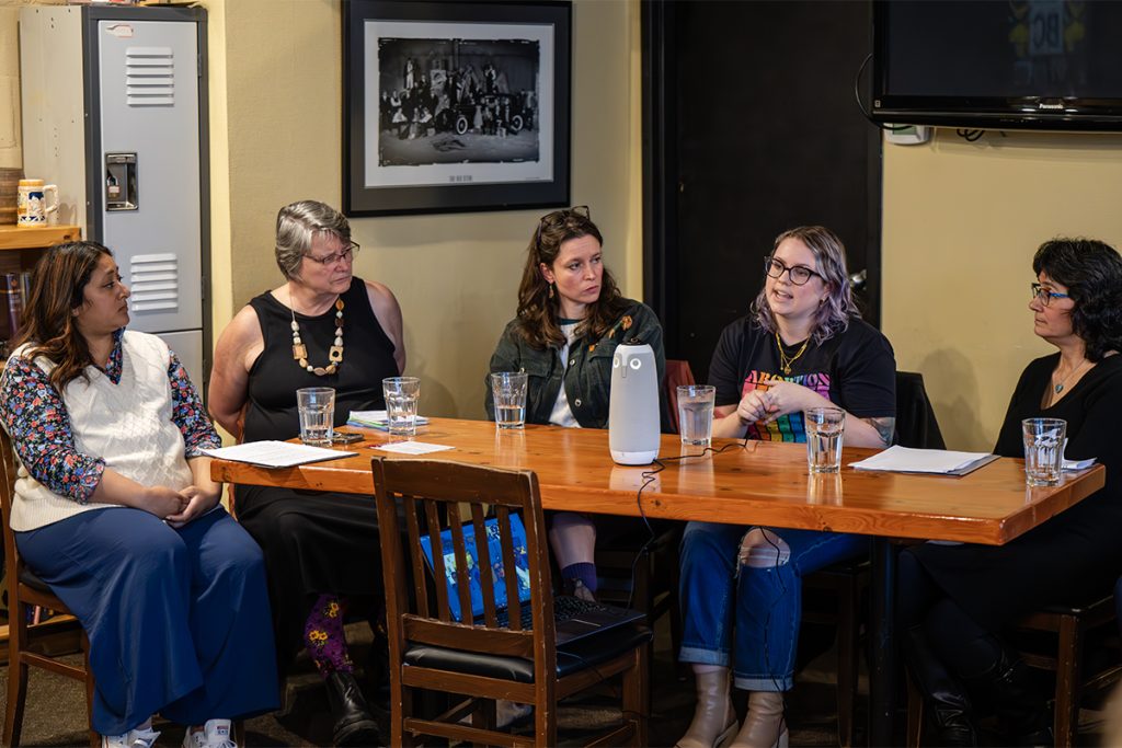 Five women sit at a table and watch one of the women speak.