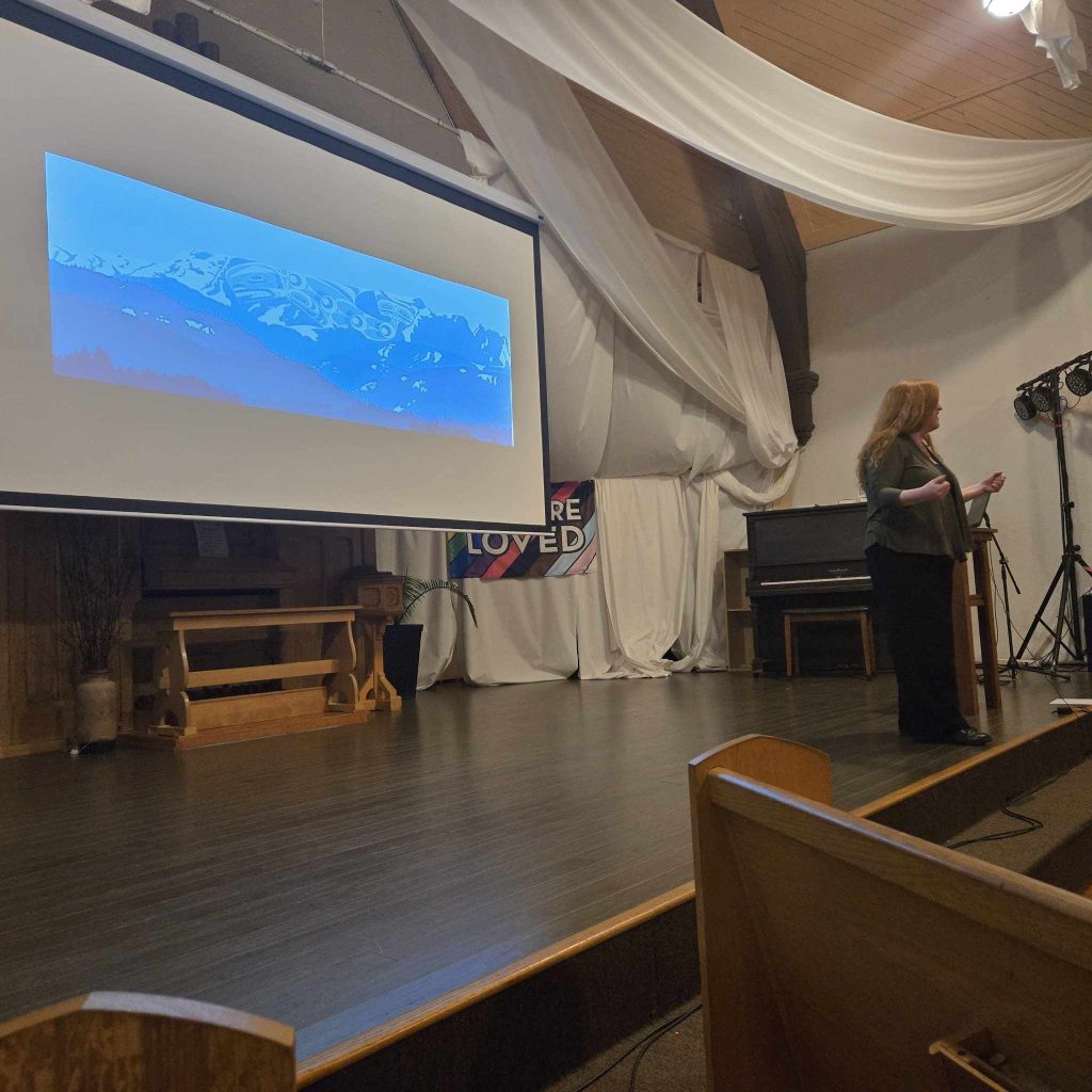A woman with long red hair stands on a small church stage, looking away from the camera and towards the audience in front of her. Behind her is a projected image of Indigenous artwork of the Comox Glacier.
