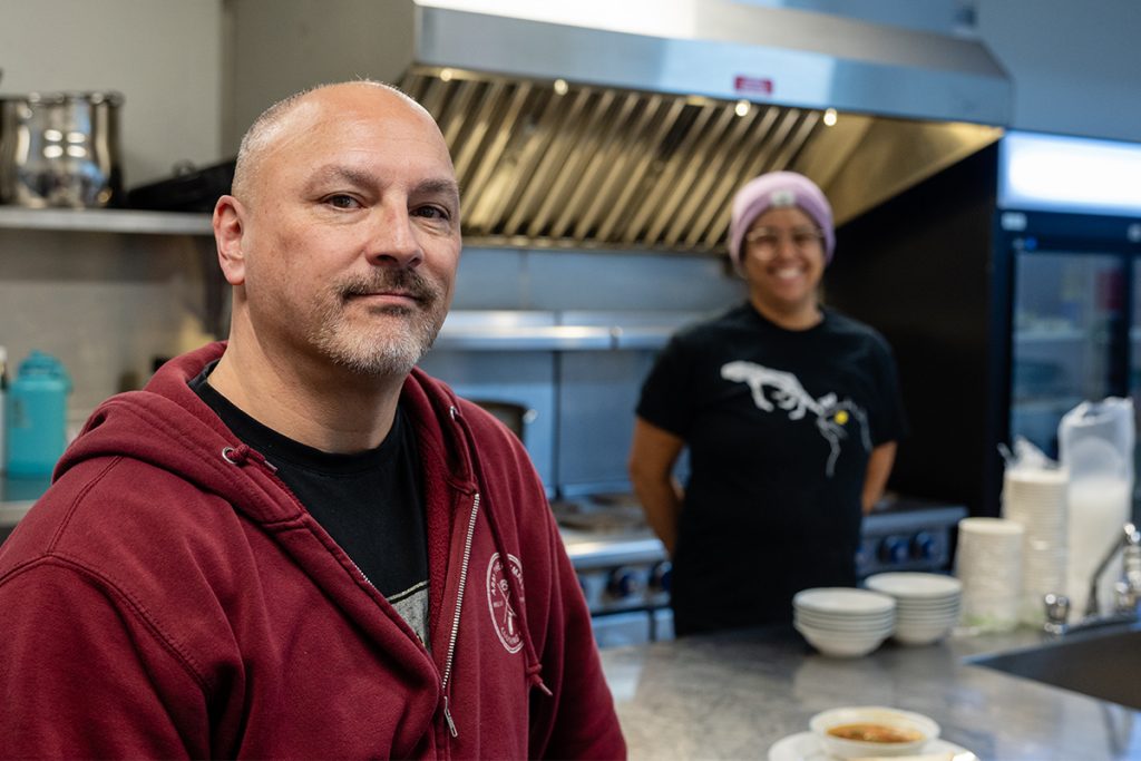 Photo of a man in a kitchen with a person in the background smiling but out of focus.