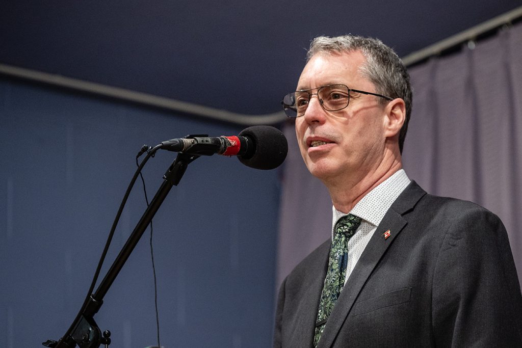 Photo of Paul Manly wearing a suit with a green tie and speaking at a microphone.