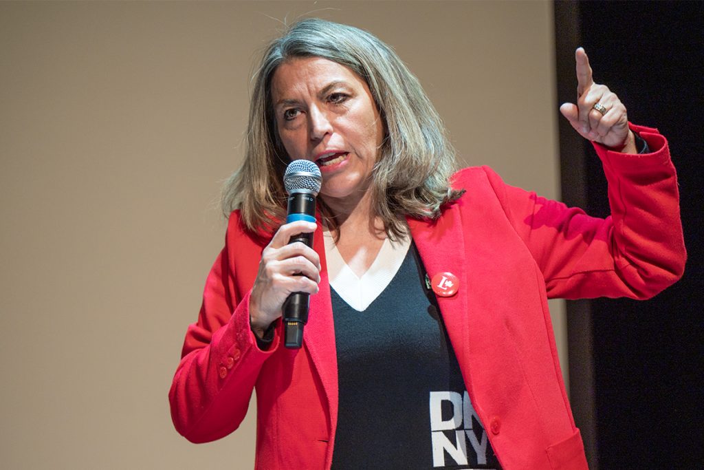 Photo of a woman wearing a red jacket and Liberal Party of Canada pin speaking into a microphone and pointing a finger in the air.