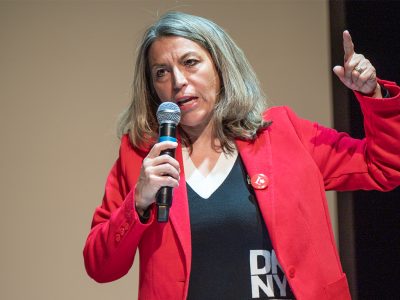 Photo of a woman wearing a red jacket and Liberal Party of Canada pin speaking into a microphone and pointing a finger in the air.