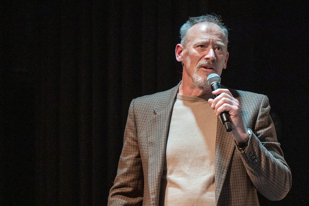 Photo of a man wearing a checkered suit jacket and sweater speaking into a microphone with a black background.
