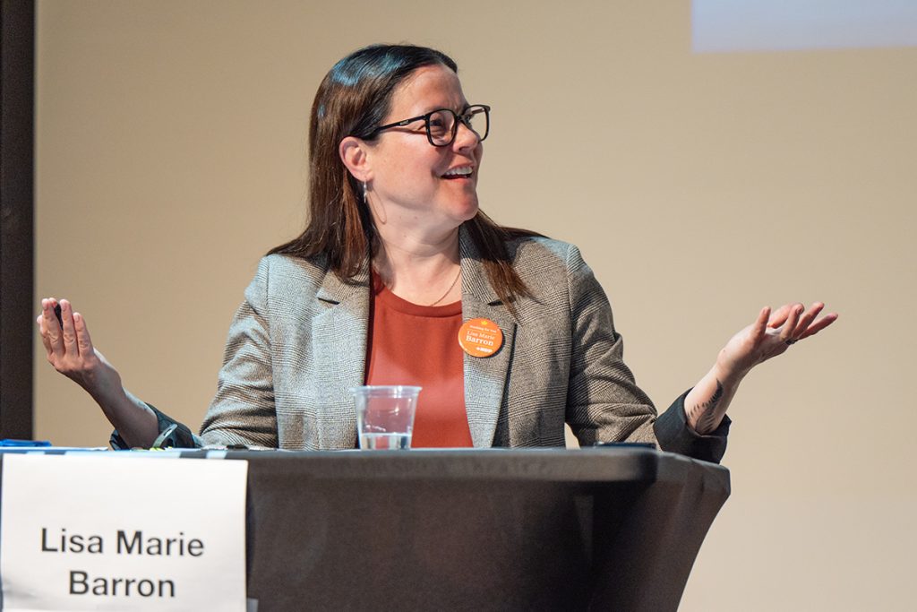Photo of a Lisa Marie Barron wearing a beige suit jacket and a red shirt with an orange NDP pin gesturing in disbelief