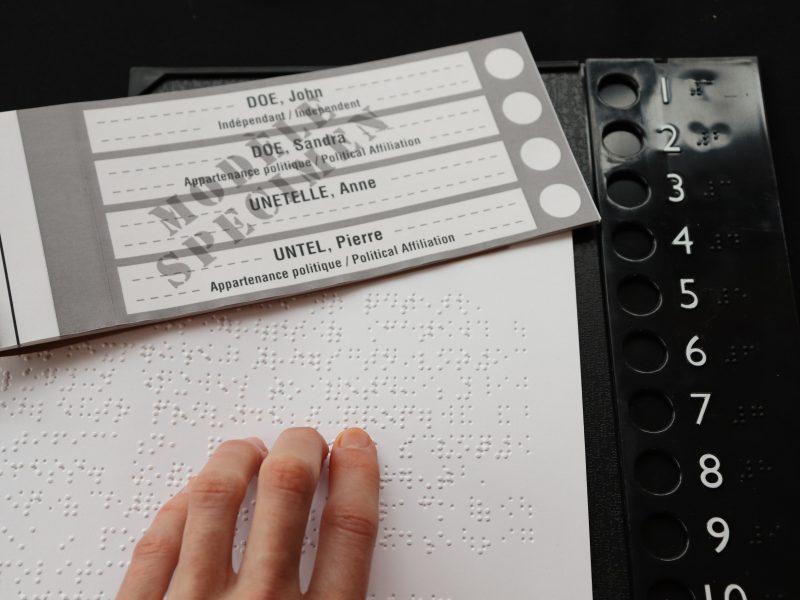 A hand on a piece of braille paper next to a blank ballot.