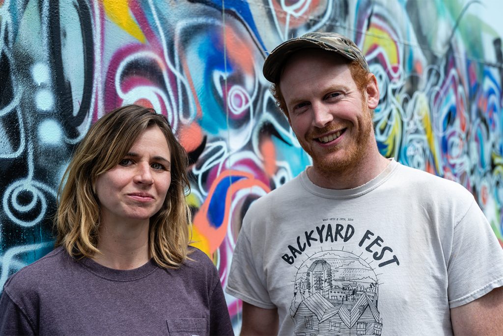 A man wearing a shirt reading Backyard Fest and a woman stand in front of a colourful graffiti mural.