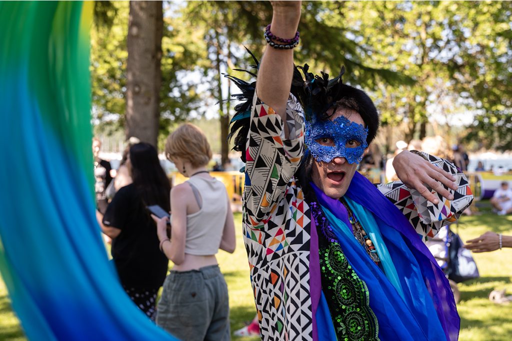A person wearing a party mask, a colourful top and a feathered hat dances while twirling a rainbow ribbon