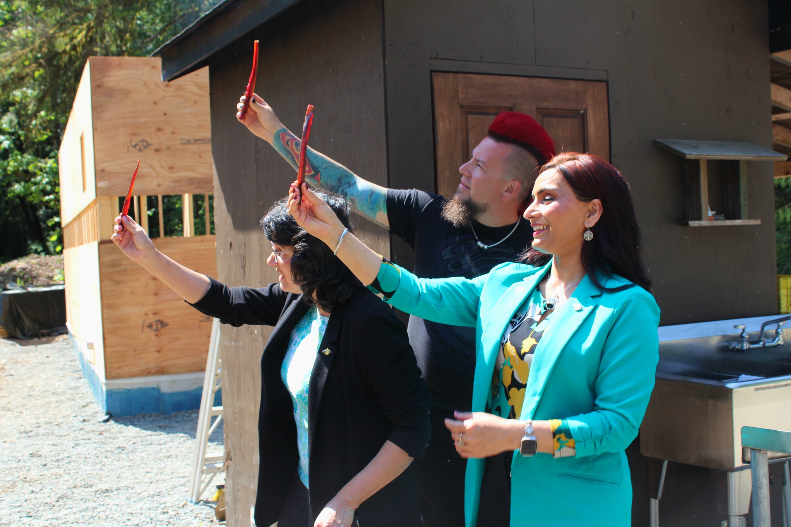 Three people are standing in front of a smokehouse holding up smoked salmon pieces, a staple of Indigenous food