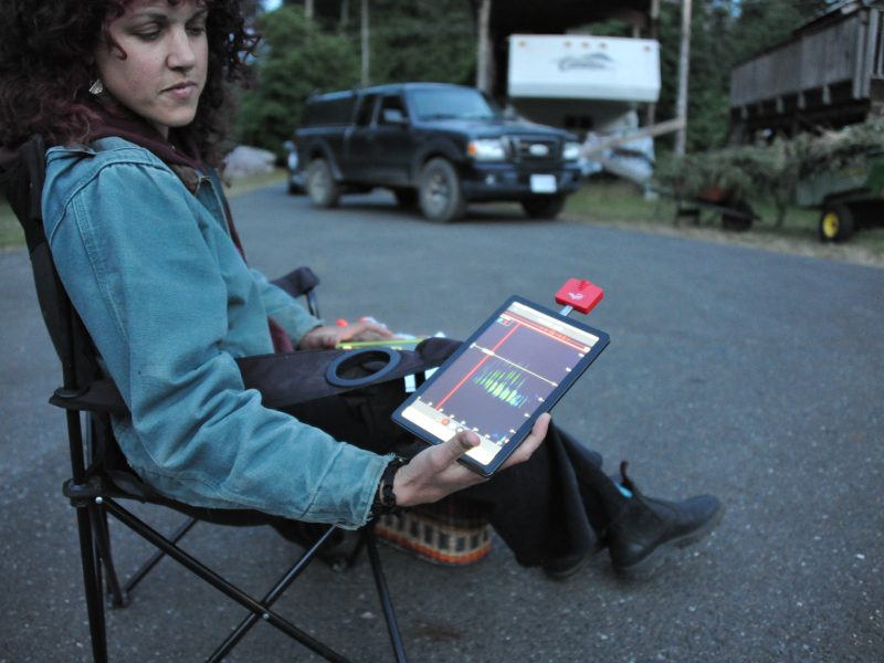 A woman sits on a camp chair on the road holding a tablet with echolocation signals.