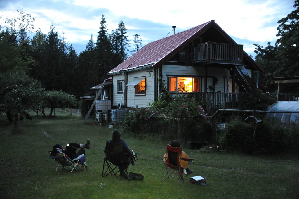 Three people sit in camp chairs in a lawn at dusk looking at the roof of a house for bats.
