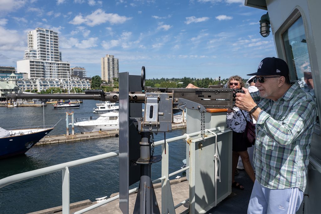 A man in civilian clothes poses with a machine gun on HMCS Nanaimo with the Nanaimo harbour in the background.