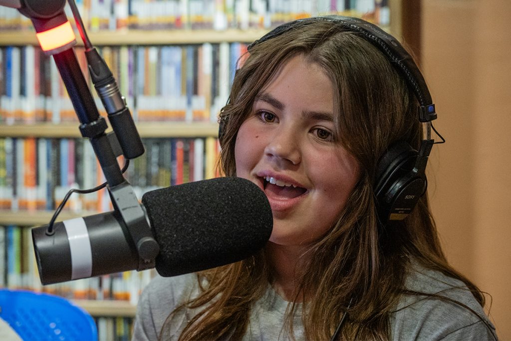 Photo of a young woman speaking into a radio microphone with a rack of CDs behind her.