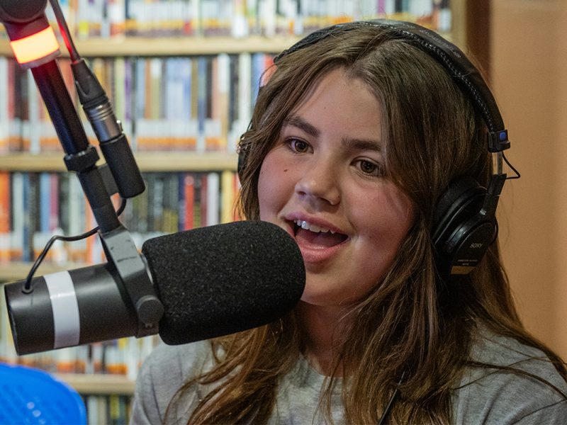 Photo of a young woman speaking into a radio microphone with a rack of CDs behind her.