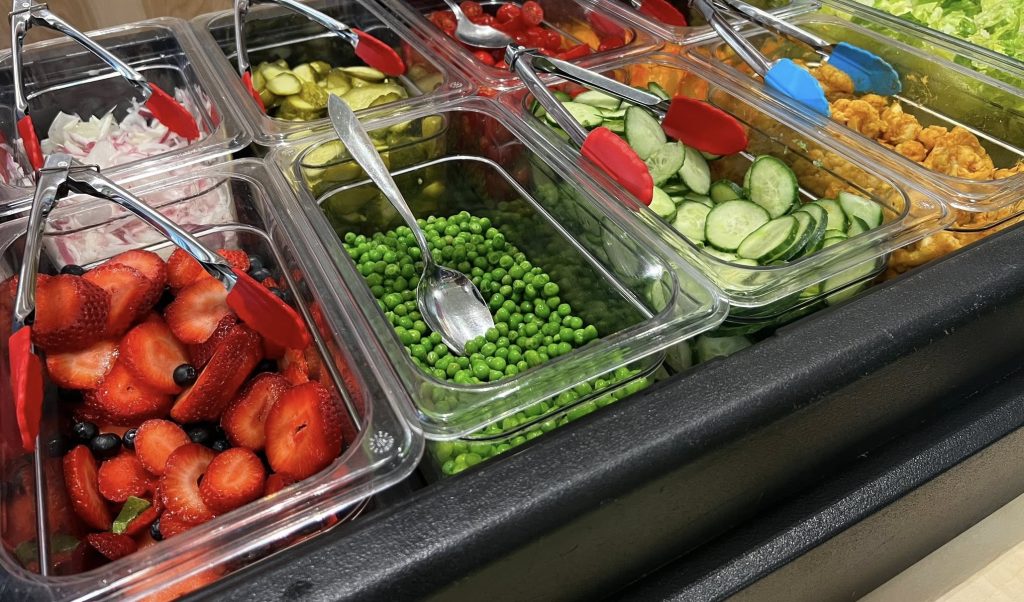 A close-up of a salad bar with strawberries and blueberries, peas, radishes, cucumbers, chickpea salad, lettuce, tomatoes, pickles and radishes. All items in a separate container with tongs.