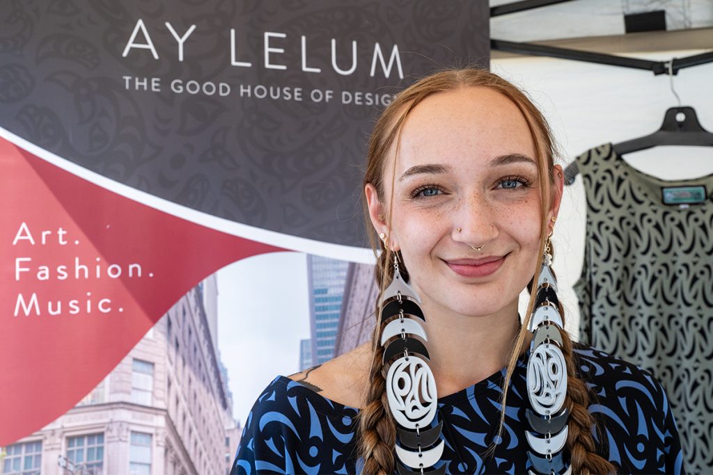 Brooklyn Chalmers wearing designer earrings stands in front of a sign for Ay Lelum The Good House of Design during Snuneymuxw Days 2025.