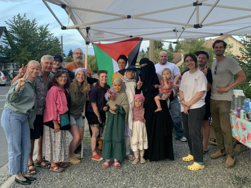 A group of people stand under a tent in front of a Palestinian flag.