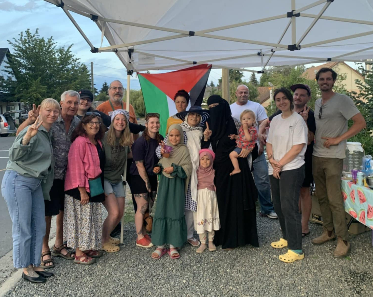 A group of people stand under a tent in front of a Palestinian flag.