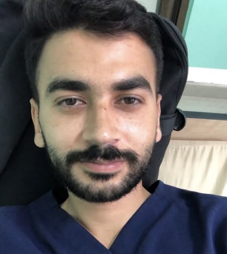 A selfie of a close-up of a man's slightly smiling face. He has a beard and short black hair, and appears to be wearing scrubs. He looks tired.
