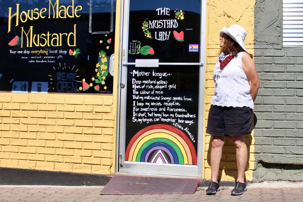 A woman stands outside a shop that is painted yellow. It says 'The Mustard Lady' on the window.