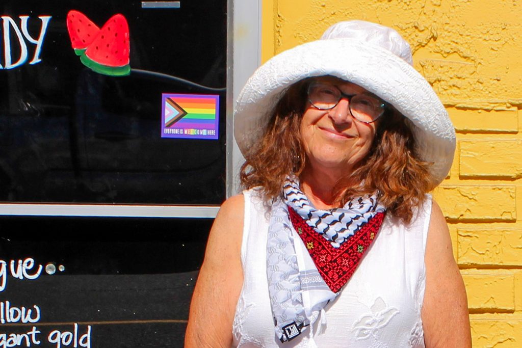 A close-up of an older woman's face. She is wearing a keffiyeh and a wide-brimmed white hat.