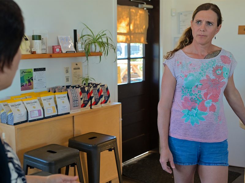 Two women in a cafe stand and look at one another cautiously.