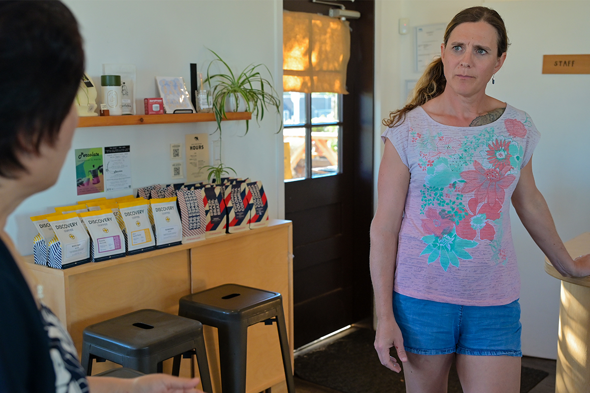 Two women in a cafe stand and look at one another cautiously.