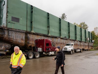 A man in high viz and a woman in a dark rain jacket walk along a beach in the rain. There is a large barge behind them.