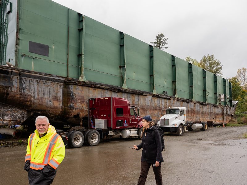 A man in high viz and a woman in a dark rain jacket walk along a beach in the rain. There is a large barge behind them.