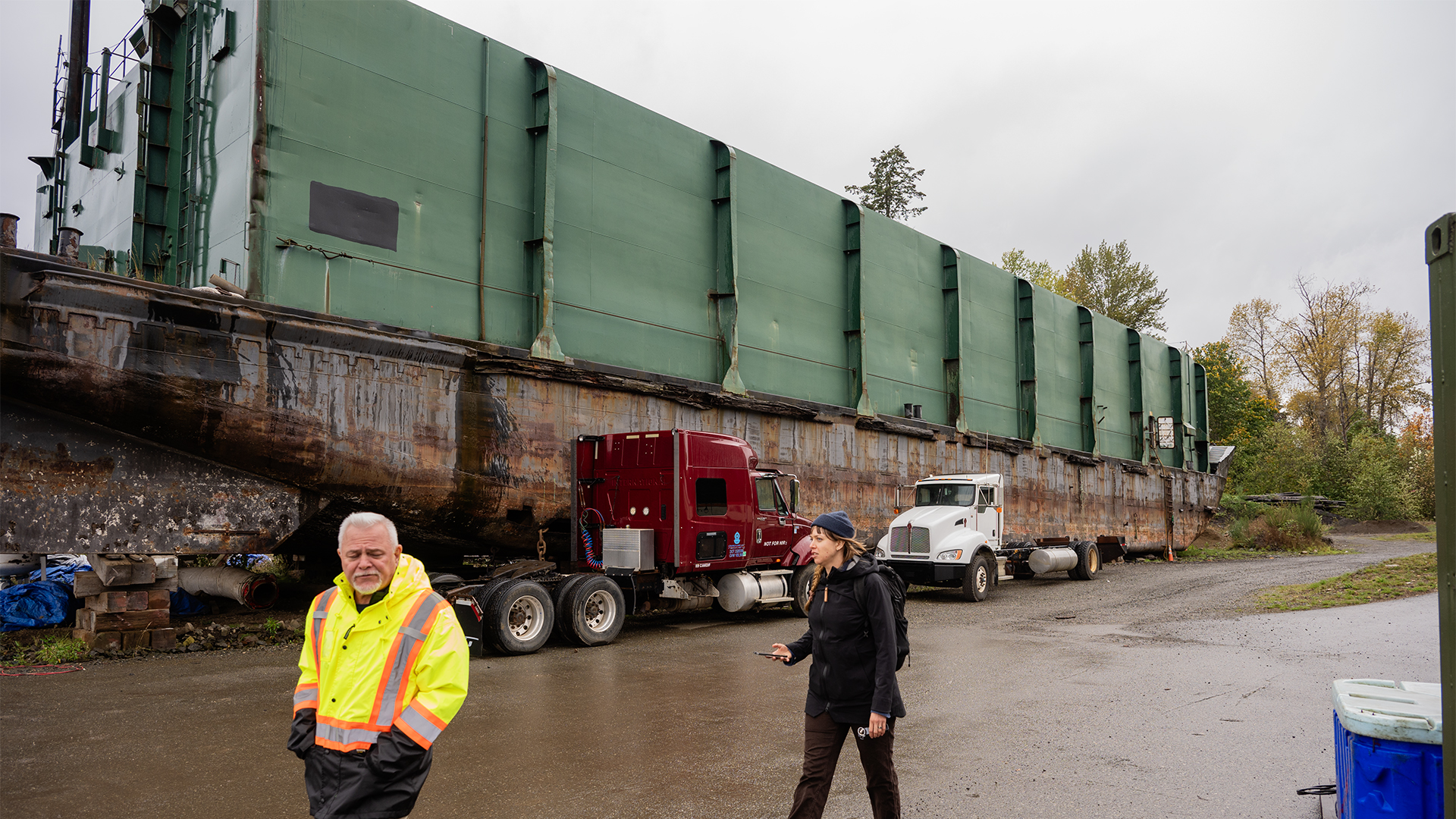 A man in high viz and a woman in a dark rain jacket walk along a beach in the rain. There is a large barge behind them.