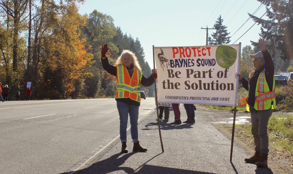 Two people stand in high viz holding a sign that says "Protect Baynes Sound Be Part of The Solution"