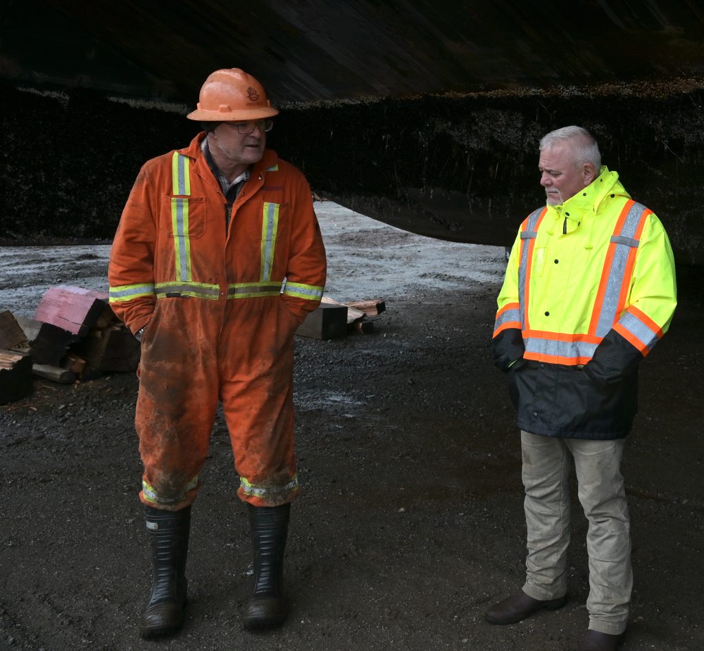 Two men in high viz stand under a shelter in the rain and chat.