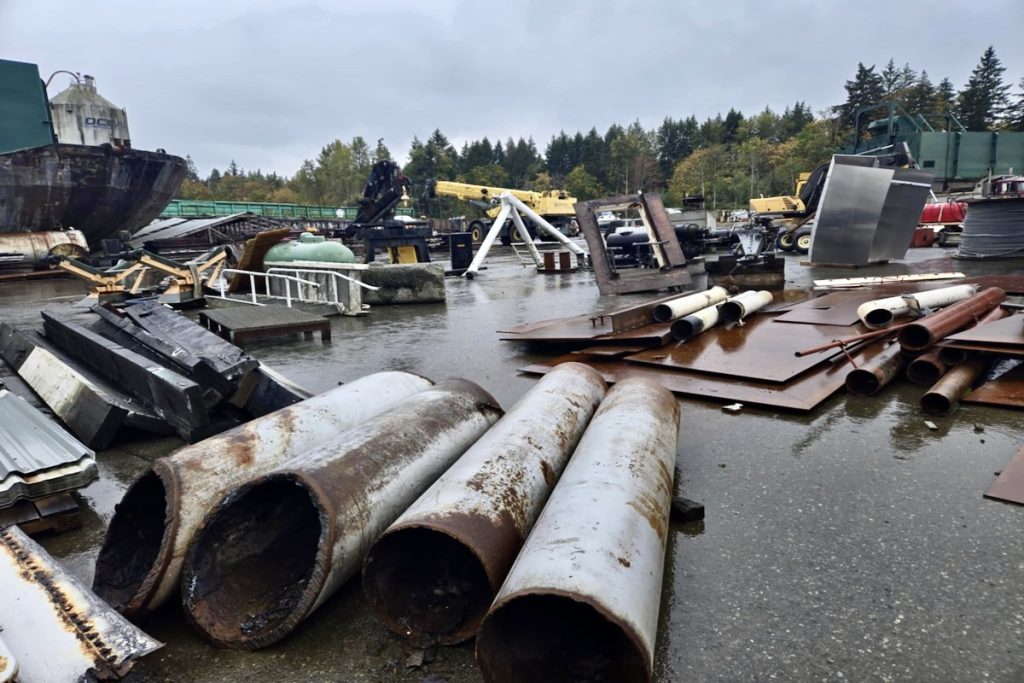 Various scrap metle laid out on what appears to be a beach, judging by part of a barge in the background. 