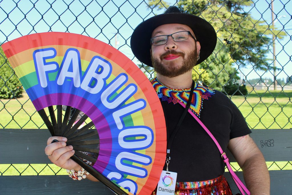 A person with a big beard and bright pink lipstick smiles and holds a large unfolded fan that says 'fabulous'