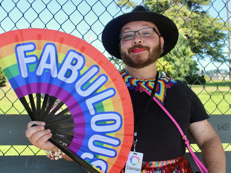 A person with a big beard and bright pink lipstick smiles and holds a large unfolded fan that says 'fabulous'