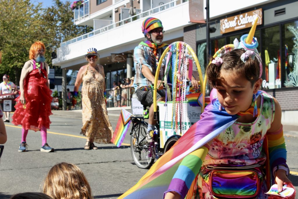 A child in rainbow colours hands out stickers along the road. A man behind her rides a bike with colourful decorations. Two people are seen walking in bright colours behind them.