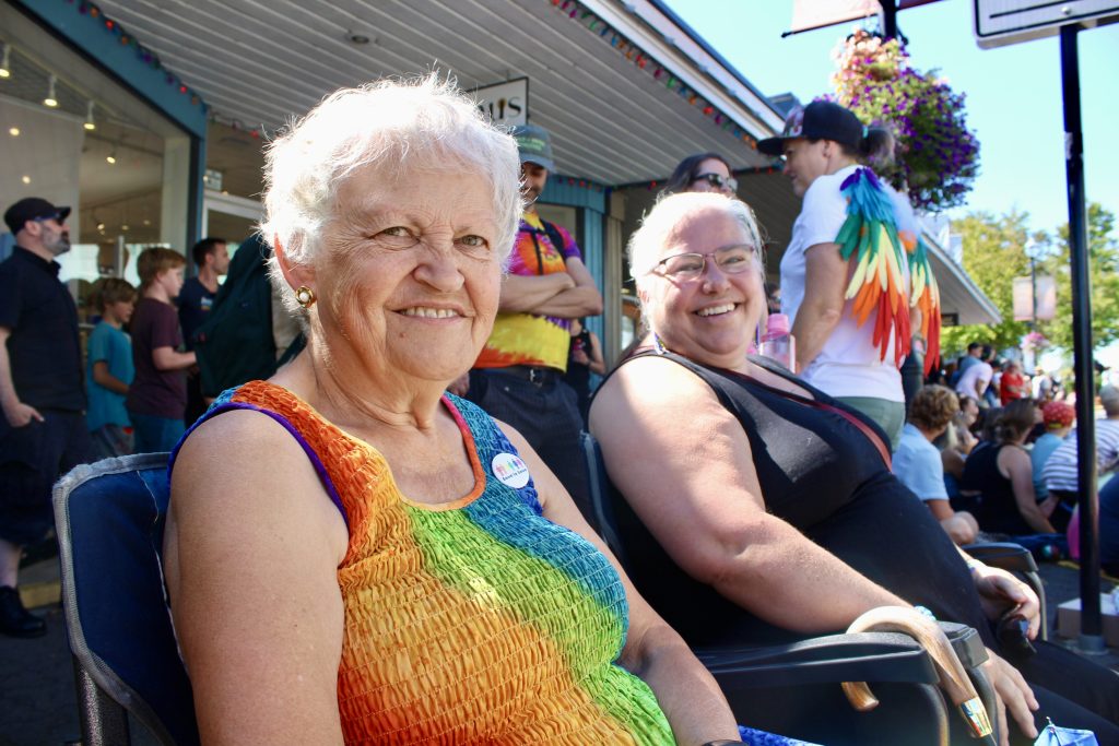 Two women watching a parade, one in bright rainbow colours, smile at the camera. One woman is an elderly woman with short hair and another is a middle-aged woman with a ponytail.