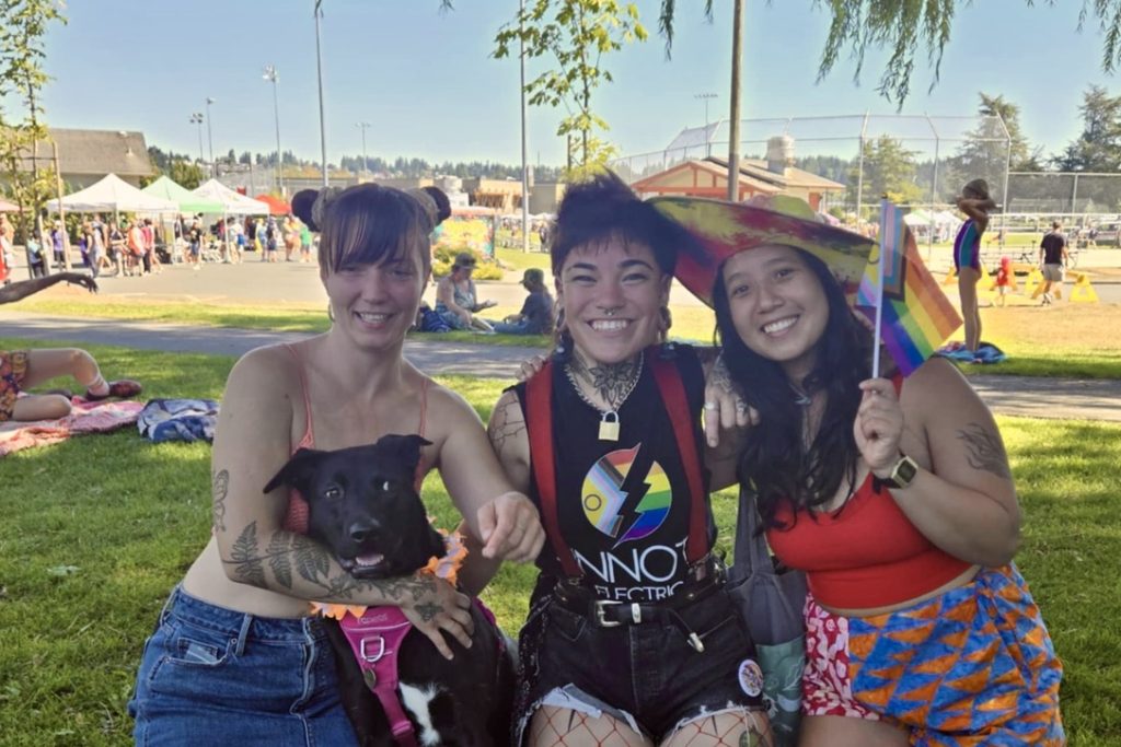Three people sitting in the grass, with a black dog. One person waves a pride flag, and another is wearing a shirt with pride colours.