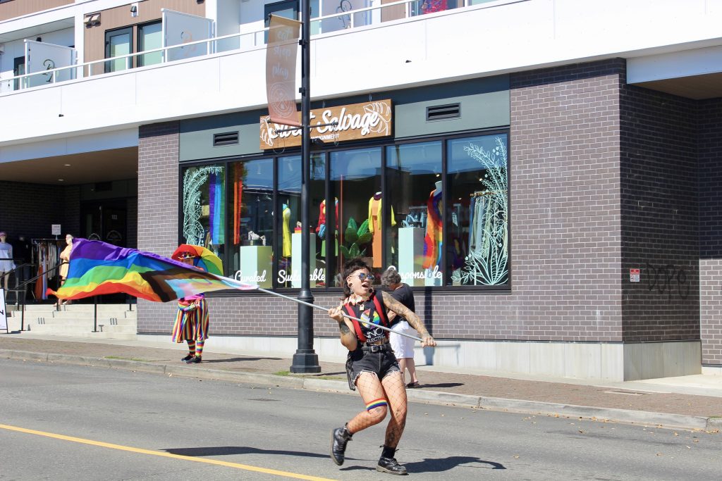 A person dances down the street waving a large pride flag. They have short dark hair and are dressed in cutoff shorts and mostly black, with a rainbow knee band and a t shirt with pride colours