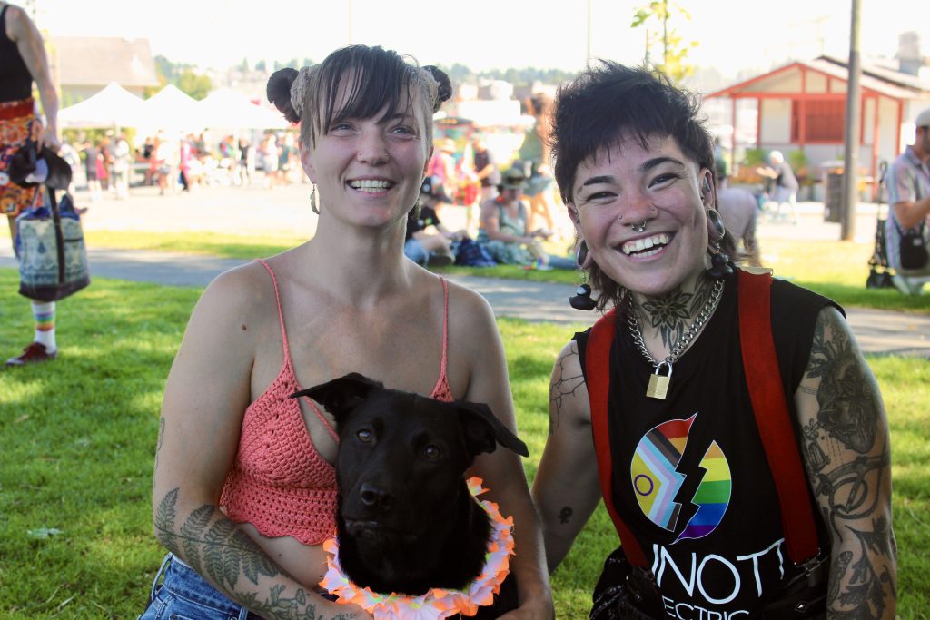 Two people sitting in the grass, with a black dog. One person is wearing a shirt with pride colours. Both are smiling