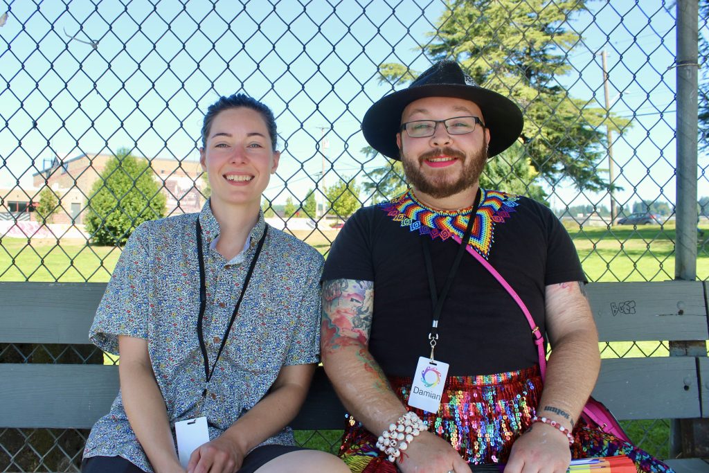 Two people, one in bright pride colours and another in a button up shirt, sit on a bench at Comox Valley Pride