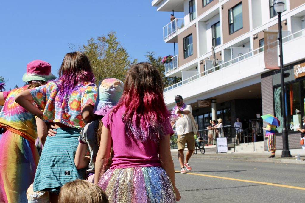photo taken from behind of children with rainbow colours and dyed purple and pink hair standing towards the street watching the Comox Valley pride parade